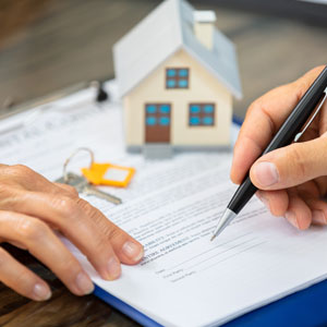 Person signing real estate documents with house model and keys on table